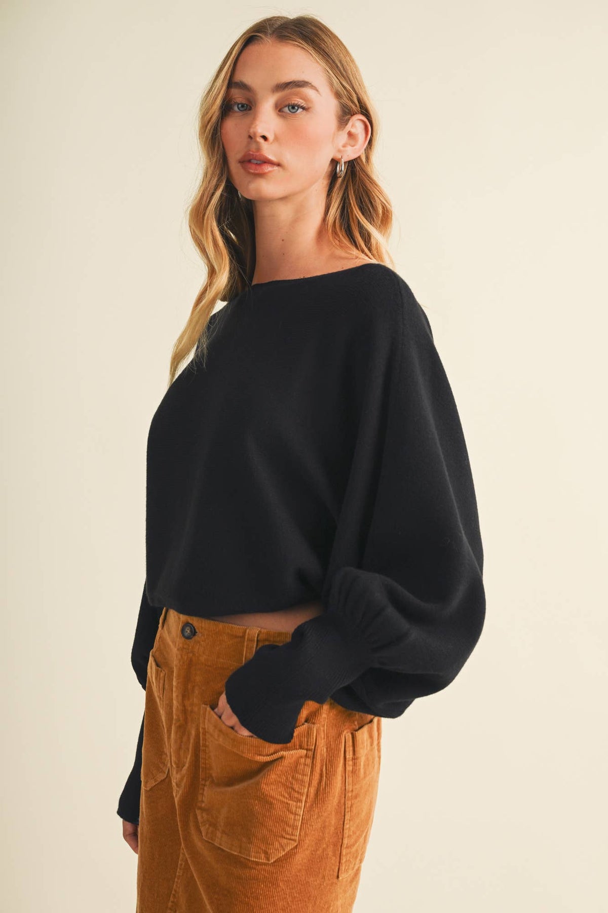 Black dolman pullover with bateau neckline and loose fit over brown pants, side view.