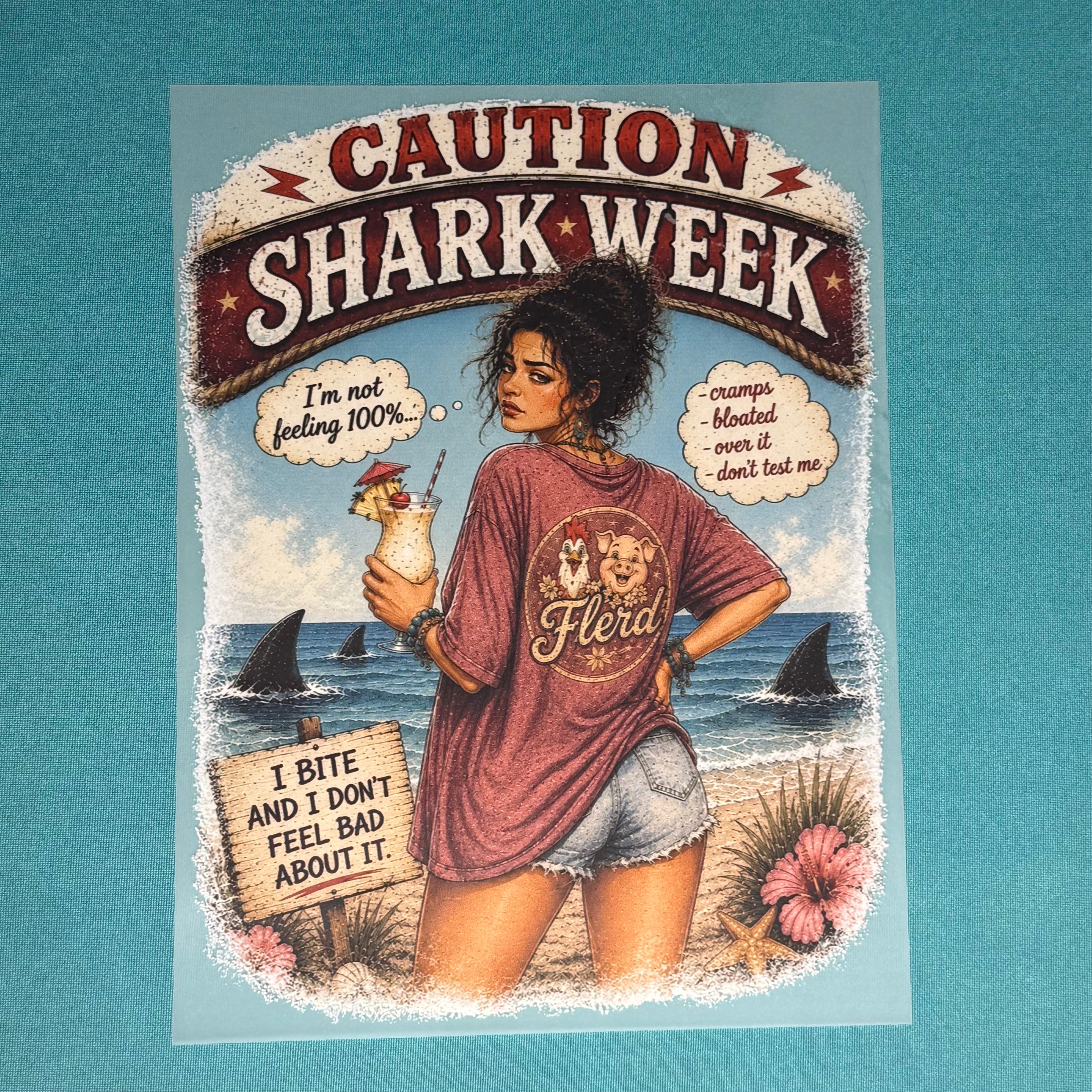 Maroon graphic T-shirt with "CAUTION SHARK WEEK" print and illustrated woman holding treat