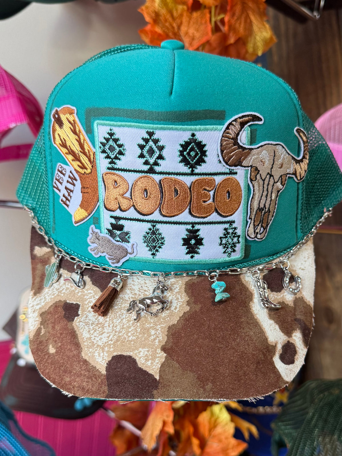 Teal rodeo trucker hat with embroidered “RODEO” patch and hanging charms at brim